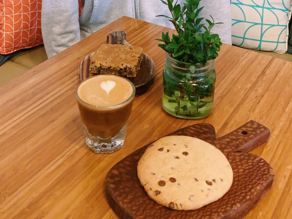 Coffee, cookie, and brownie on wood table and cutting boards with plant on the table.