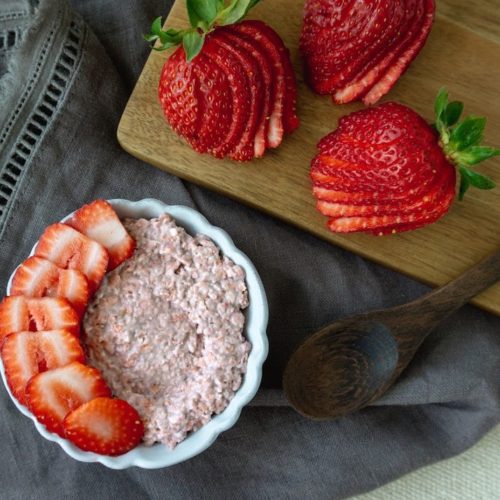 Strawberry Creme Overnight Oats topped with fresh strawberries on a gray cloth, next to a cutting board with more fresh strawberries.