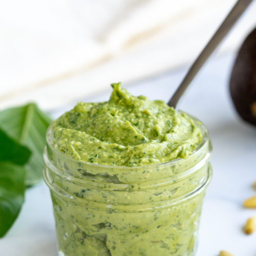 Avocado pesto in a mason jar with a spoon on a white marble top along with fresh basil and avocado