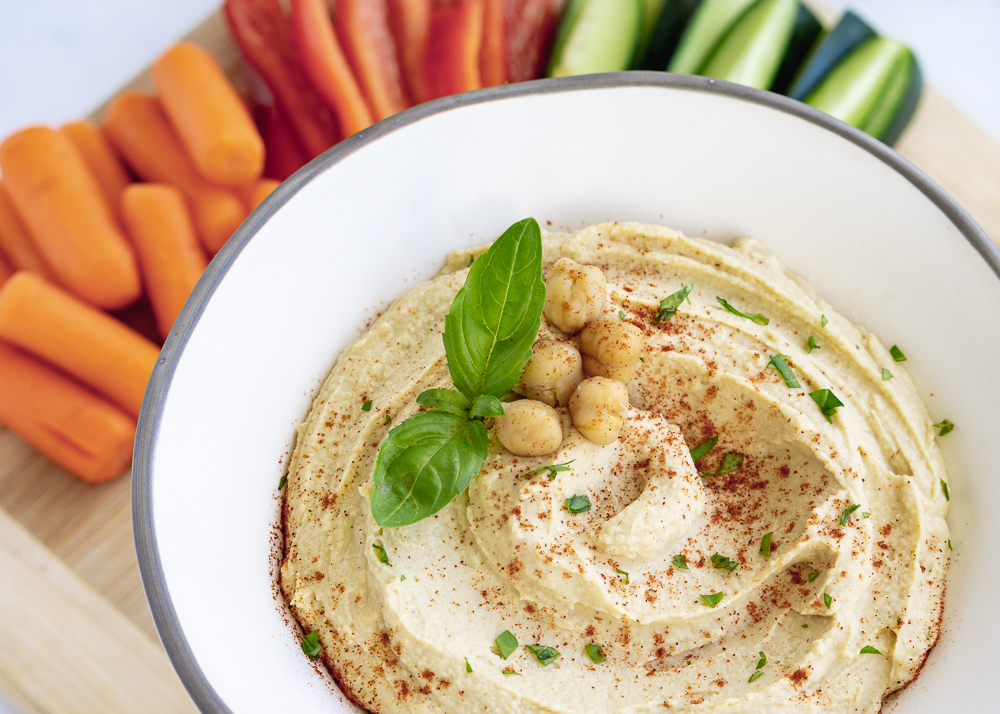 Hummus in a white bowl surrounded by baby carrots, red bell pepper slices, and cut up cucumber.
