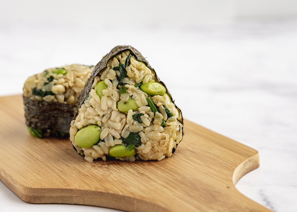 Two edamame rice musubis on a wood cutting board against a marble background.