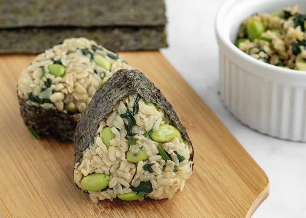 Two rice musubis on a wood cutting board with edamame rice and seaweed sheets in the background.