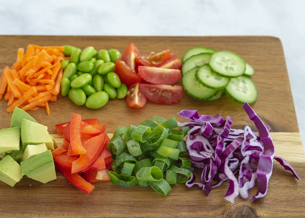 Chopped cucumbers, purple cabbage, bell pepper, edamame, carrots, avocado, tomatoes, and green onion on a wood cutting board.