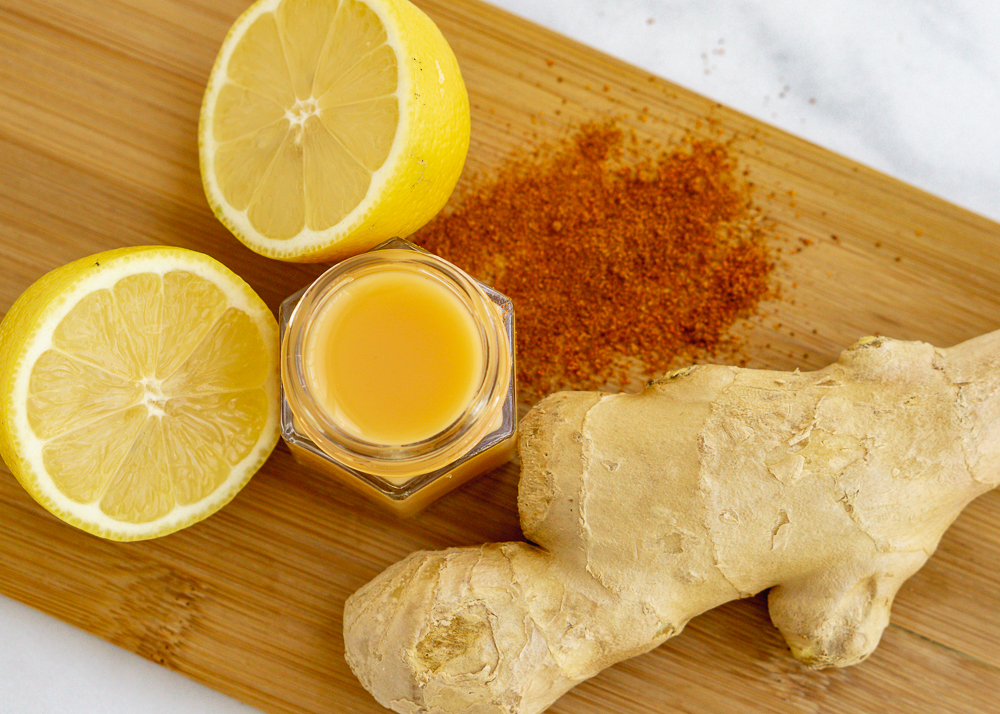 Lemon ginger wellness shot on a wood cutting board surrounded by two lemon halves, 1/2tsp cayenne pepper, and ginger root.