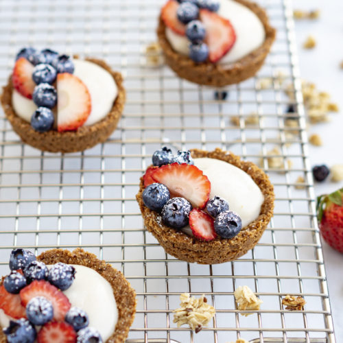 Six vegan fruit tarts on a cooling rack with granola, strawberry, and blueberries around them.