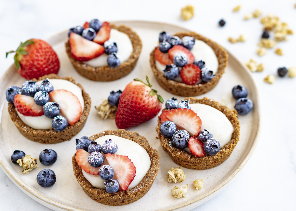 Five vegan fruit tarts on a cream plate with granola, blueberries, and strawberries.