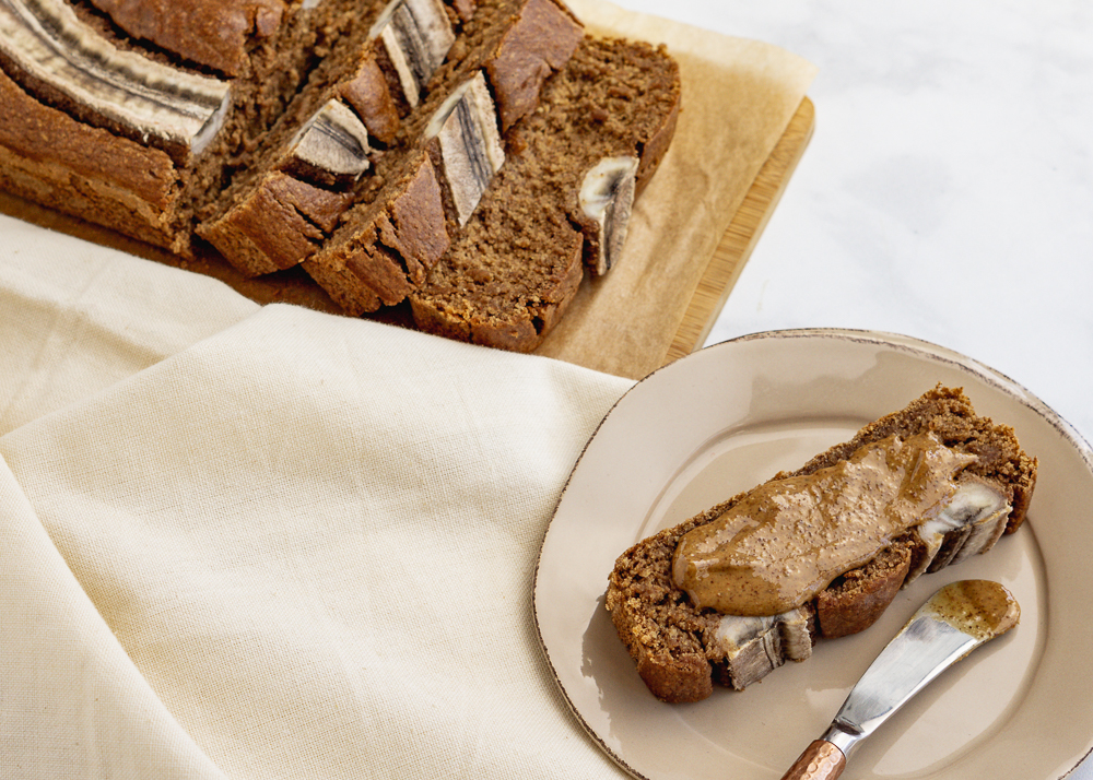 Cut banana bread loaf on a wood cutting board, next to a slice of banana bread on a plate topped with almond butter