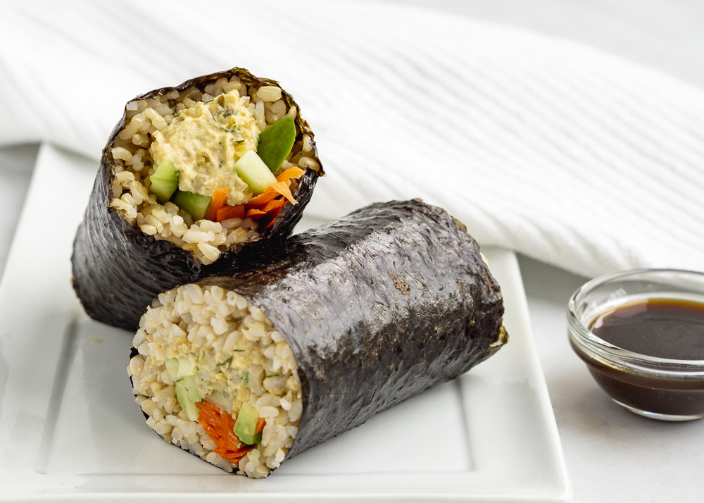 Two vegan sushi burritos on a white plate with a white napkin and soy sauce in a small bowl