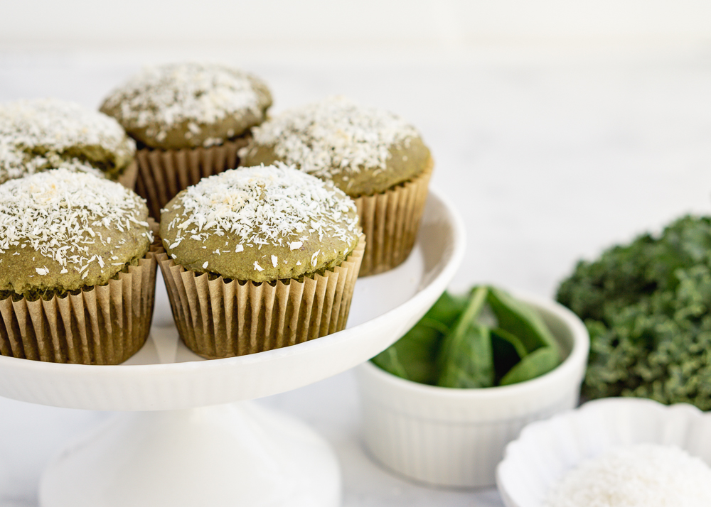 Five green juice pulp muffins on a white cake stand with kale and spinach in the background in white bowls.