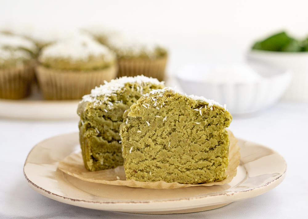 Green juice pulp muffin cut in half on a beige dish with five whole muffins in the background on a white plate