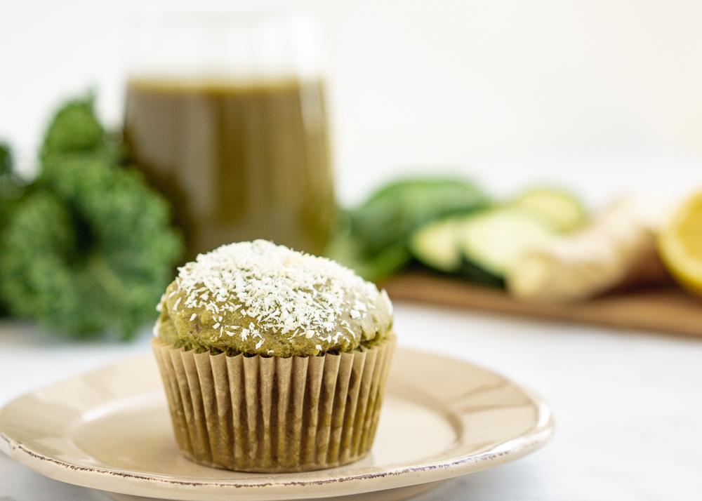 Single green juice pulp muffin on a beige plate with leafy green, ginger, and a glass of green juice in the background