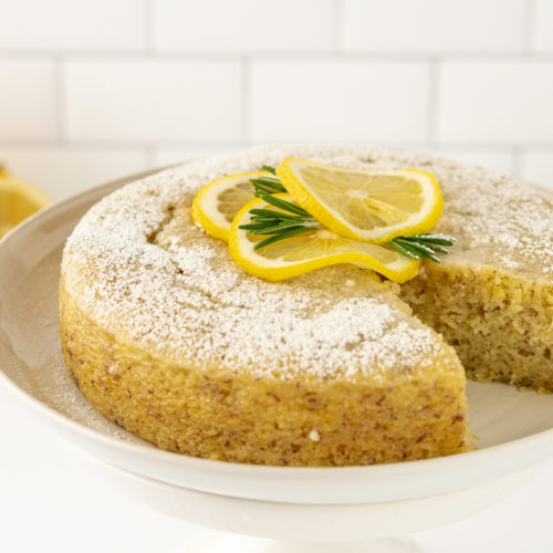 Vegan lemon cake topped with powdered sugar, lemon, and rosemary on a white plate with white tiles and lemon in the background.