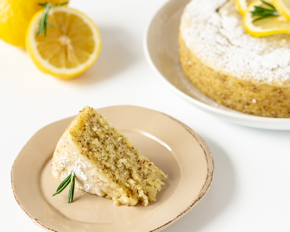 Slice of vegan lemon cake on a brown plate with lemon and lemon cake in the background