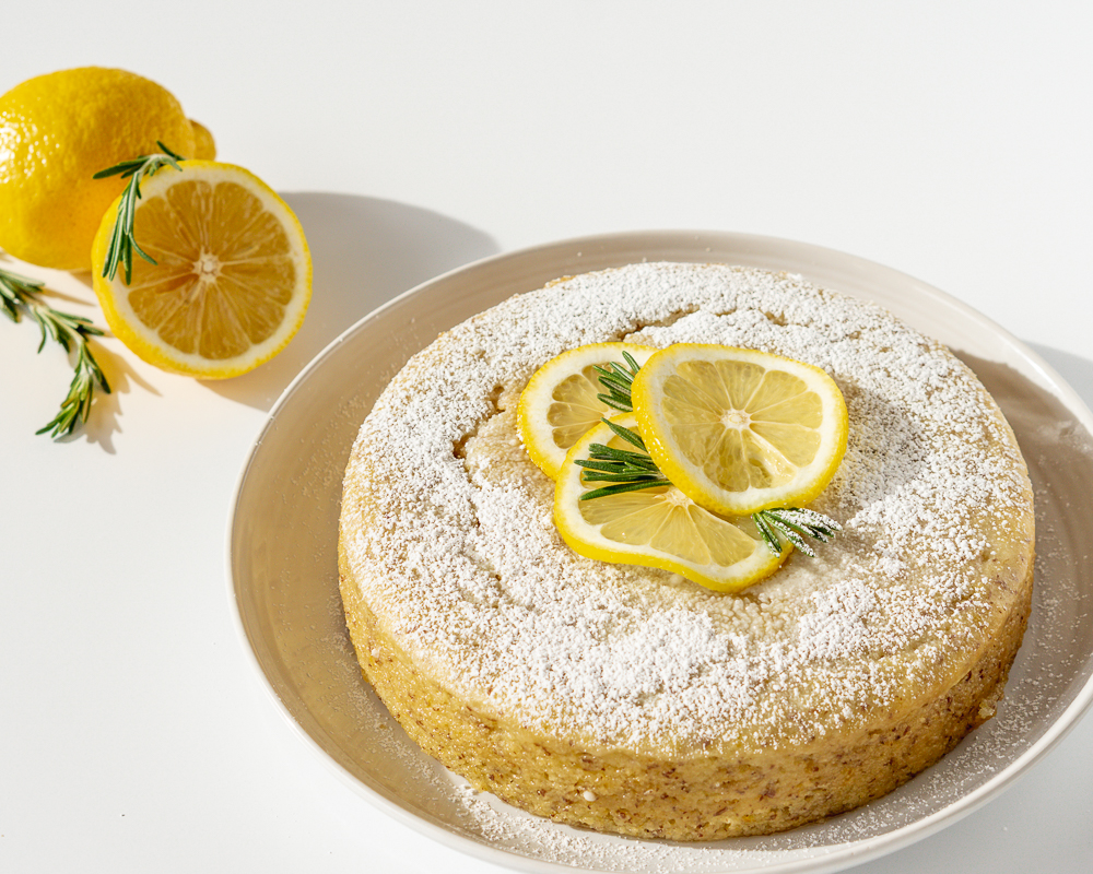 Vegan lemon cake on a white plate with lemon halves and rosemary in the background