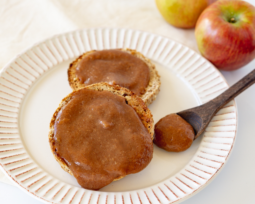 Homemade apple butter spread on an English muffin with a wood spoon and fresh apples in the background.